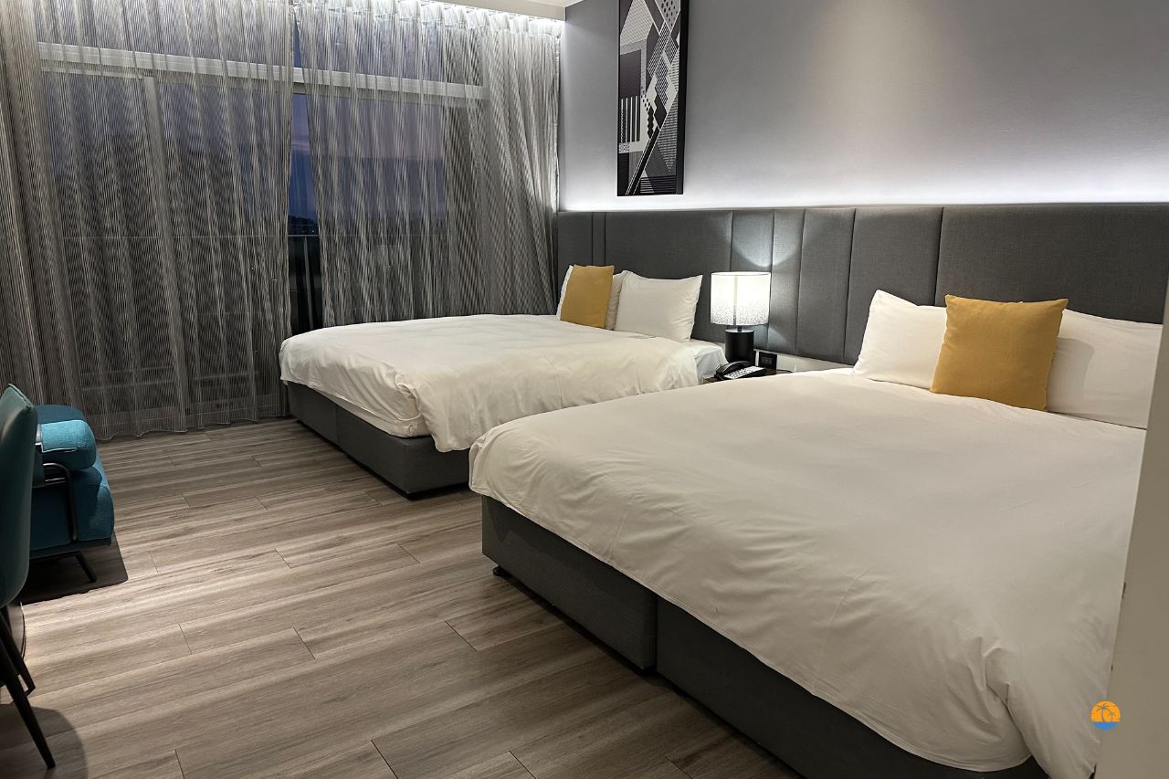 Pokój hotelowy z dwoma łóżkami, jasną podłogą i dużym oknem z zasłonami